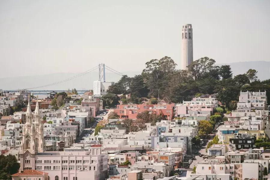 Abgebildet ist der Coit Tower in San Francisco mit der Bay Bridge im Hintergrund und einem mit Häusern bedeckten Hügel im Vordergrund.